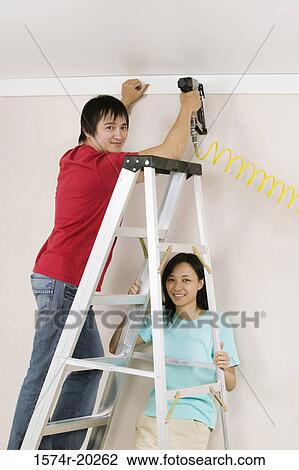 Portrait of a young man using a drill on a wall with a young woman holding a ladder View Large Photo Image Stock Image - Portrait of a young man using a drill on a wall with a young woman holding a ladder. Fotosearch