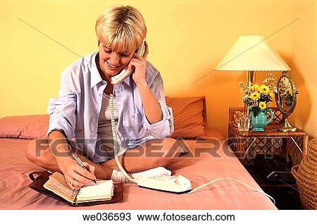 Stock Image - Young blond woman sitting cross legged on her bed and talking on the landline telephone whilst making appointments in her diary. Fotosearch