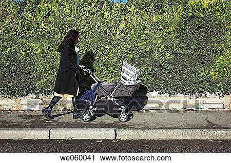Stock Image - Young woman pushing a pram walking through a park.. Fotosearch