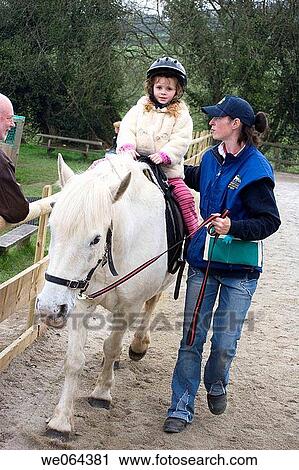 Stock Image - 4 year old girl riding a horse at a farm, with the assistant holding her. Fotosearch