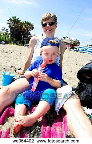 Stock Image - wide angle of 3 year old boy sitting between his mothers legs, on the beach, eating a lolly. Fotosearch