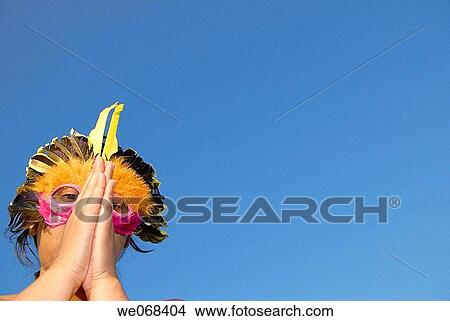 Picture - Woman wearing a colored mask resembling a bird. Hands joined as if worshiping-praying. Fotosearch