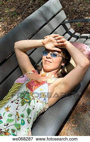 Stock Photo - Young woman, wearing sunglasses, lying on a bench at a park. Fotosearch