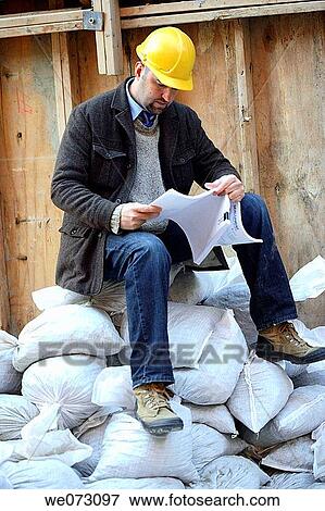 Architect, Engineer, General Contractor with hard hat sitting on sand bags on construction site reading plans View Large Photo Image Stock Photo - Architect, Engineer, General Contractor with hard hat sitting on sand bags on construction site reading plans. Fotosearch