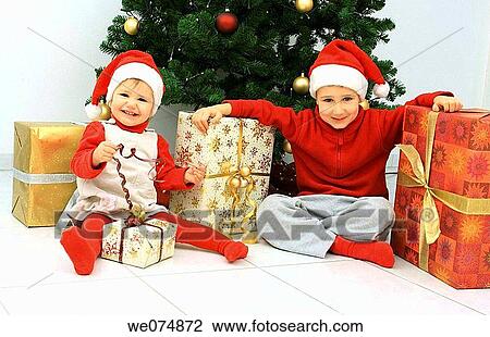 Stock Image - Child wearing Father Christmas hat with presents in front of Christmas Tree. Fotosearch