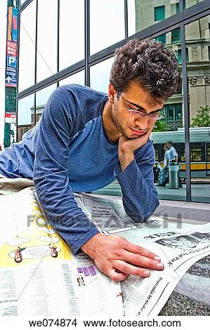 Picture - Male college student, wearing glasses, sitting outside next to a city building, reading a newspaper.. Fotosearch