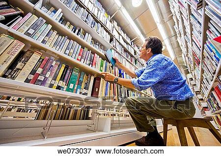 Man sitting, holding a book, and looking through a bookshelf in a library. View Large Photo Image Stock Photo - Man sitting, holding a book, and looking through a bookshelf in a library.. Fotosearch