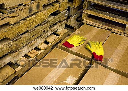Stock Image - Pair of yellow gloves laying on top of cardboard boxes, which are next to wood pallets.. Fotosearch
