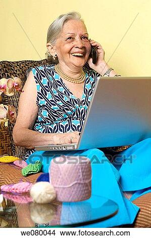 Picture - Senior lady speaking on a cell phone using a laptop computer. Fotosearch