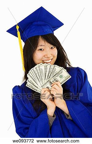 Stock Image - An Asian teenage in blue graduation gown and smiling while hold US money to illustrate to high cost of education She is on a white background. Fotosearch