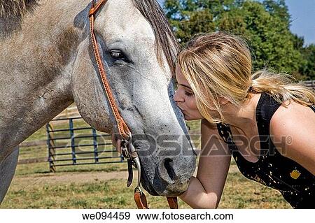 Stock Photo - Blonde woman kissing a horse on the nose the is standing in a pasture. Fotosearch