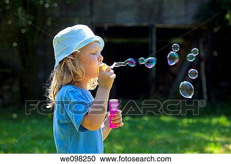 Stock Image - boy with long blond hair blowing soap bubbles outdoors. Fotosearch