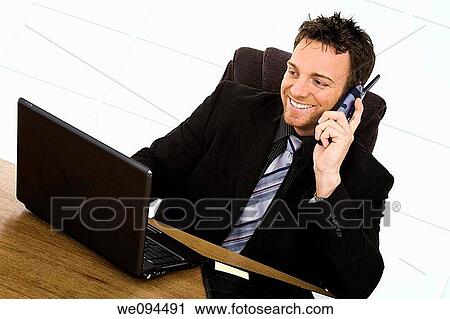 Caucasian businessman setting at a desk talking on the phone and smiling while working on a laptop View Large Photo Image Stock Image - Caucasian businessman setting at a desk talking on the phone and smiling while working on a laptop. Fotosearch