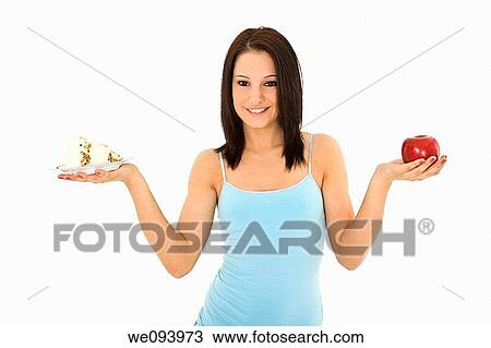 Stock Image - Caucasian woman holding an apple and slice of cake trying to decide which one to eat. Fotosearch