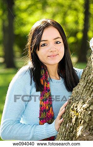 Stock Image - Caucasion teenage girl looking out from behind a tree and smiling. Fotosearch