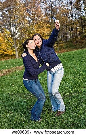 Stock Photo - Two caucasian teenage females horsing around in a park using a cell phone to take their pictures. Fotosearch