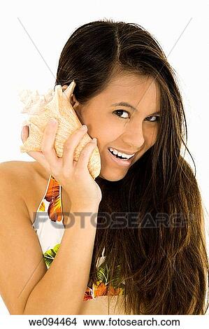 Picture - Young Asian woman holding a Conch Shell to her ear to listen to the ocean and smiling. Fotosearch