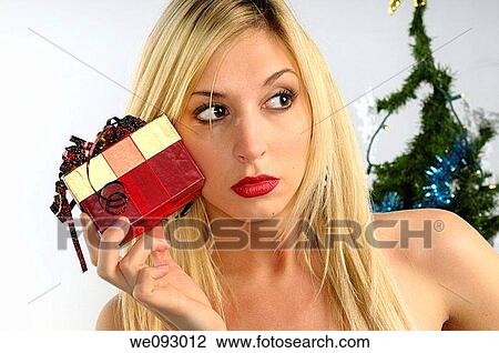 Young blond woman holding a Christmas present up to her cheek by a Christmas tree View Large Photo Image Stock Image - Young blond woman holding a Christmas present up to her cheek by a Christmas tree. Fotosearch