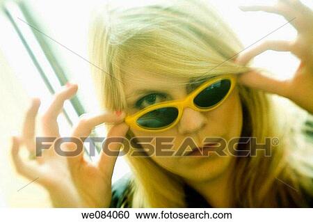Stock Image - Young blond woman, wearing sunglasses, standing by her living room window. Fotosearch