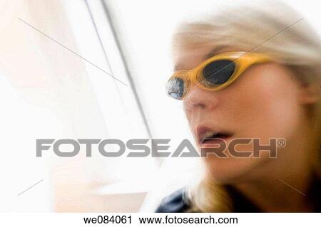 Stock Image - Young blond woman, wearing sunglasses, standing by her living room window. Fotosearch