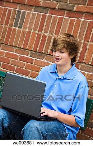 Young teenage boyl setting down on a bench and working on a laptop computer outside a school View Large Photo Image Stock Image - Young teenage boyl setting down on a bench and working on a laptop computer outside a school. Fotosearch