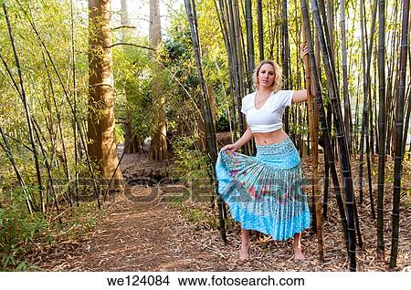 Picture - A happy 22 year old blond woman wearing a skirt and white top walking among bamboo canes in a garden.. Fotosearch