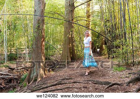 Stock Image - A happy 22 year old blond woman wearing a skirt and white top walking among bamboo canes in a garden.. Fotosearch