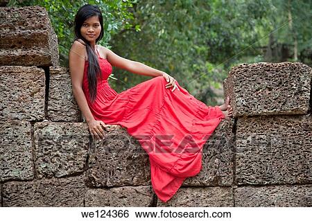 Beautiful Cambodian woman wearing a red dress sitting on a wall in Ta Phrom temple in Angkor Cambodia. View Large Photo Image Stock Photograph - Beautiful Cambodian woman wearing a red dress sitting on a wall in Ta Phrom temple in Angkor Cambodia.. Fotosearch