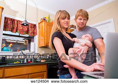 Picture - Young Parents with baby looking at laptop computer screen in their kitchen.. Fotosearch