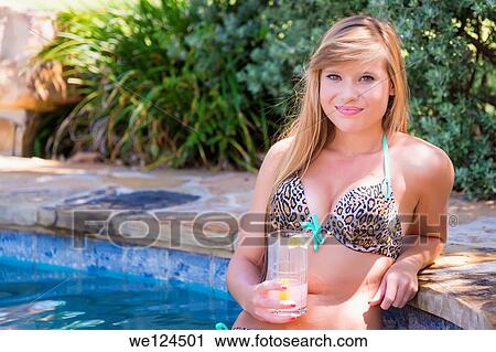 Stock Image - Young woman in leopard bikini, Caucasian, 18 years, next to domestic swimming pool area, holding beverage glass.. Fotosearch