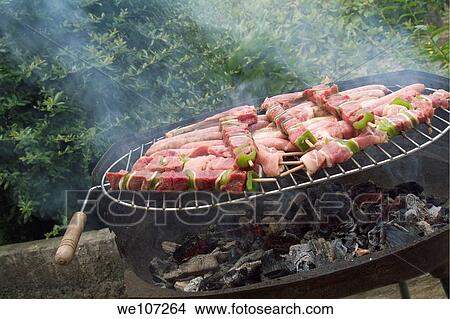 Picture - Sausages and meat on skewers grilling on a barbeque outside. Fotosearch