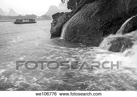 Stock Photograph - Waterfall cascading over rocks with a boat on Li Jiang River in the background, Yangshuo, Guangxi, China. Fotosearch