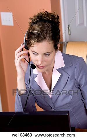 Stock Photo - Woman in early 20s working on computer and talking on headset telephone at home. Fotosearch