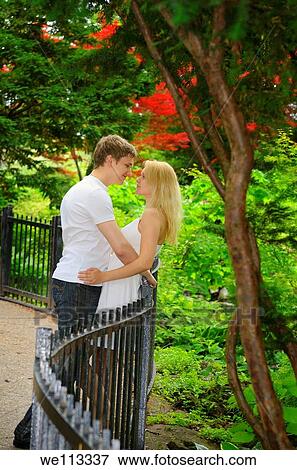 Stock Photo - Couple in love leaning on a fence for a path in a park forest. Fotosearch