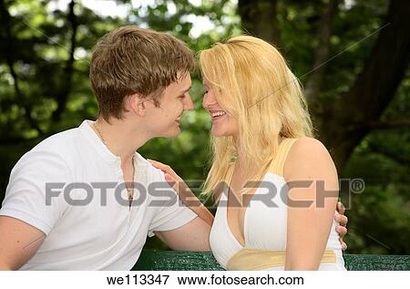 Stock Photo - Lovers touching and smiling while sitting on a park bench. Fotosearch