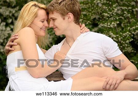 Loving couple touching and smiling while lounging on a bench in a park with flower blossoms View Large Photo Image Stock Photograph - Loving couple touching and smiling while lounging on a bench in a park with flower blossoms. Fotosearch