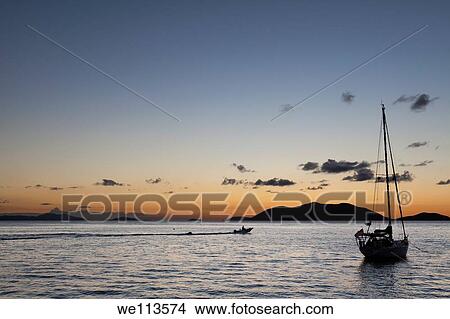 Silhouette of sailboat moored in water and power boat riding by in the orange glow of sunset in the Caribbean View Large Photo Image Picture - Silhouette of sailboat moored in water and power boat riding by in the orange glow of sunset in the Caribbean. Fotosearch