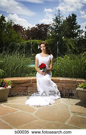 Stock Photography - A young modern bride sitting on a wall and holding a bunch of flowers. Fotosearch