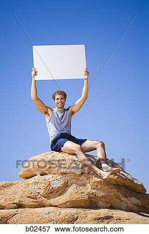 Stock Photo - Low angle view of a young man sitting on a rock with a blank sign holding up. Fotosearch