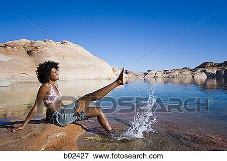 Stock Photo - Profile of a young woman sitting on a rock and splashing water. Fotosearch