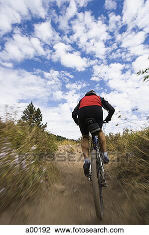 Stock Image - Rear view of a mature man mountain biking on a dirt road. Fotosearch