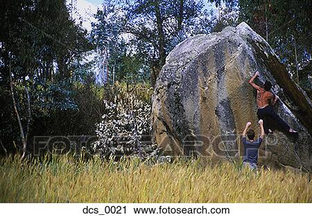 Stock Image - Rear view of two men climbing a boulder. Fotosearch
