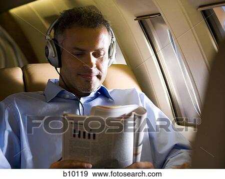 A businessman listening to music on headphones and reading a magazine in an airplane View Large Photo Image Stock Photo - A businessman listening to music on headphones and reading a magazine in an airplane. Fotosearch