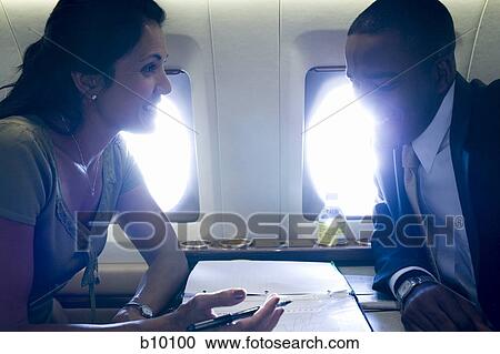 Stock Image - A businesswoman and a businessman sitting face to face in an airplane and talking. Fotosearch