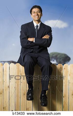 Stock Image - Businessman sitting on fence smiling. Fotosearch