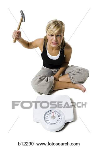 Stock Image - Frustrated dieting woman smashing bathroom scale with a hammer. Fotosearch