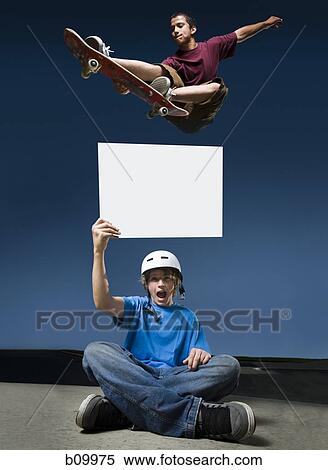 Low angle view of a teenage boy jumping with a skateboard over another teenage boy holding a blank sign View Large Photo Image Stock Photography - Low angle view of a teenage boy jumping with a skateboard over another teenage boy holding a blank sign. Fotosearch
