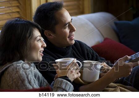 Stock Photo - Man and woman on sofa with mugs and remote. Fotosearch