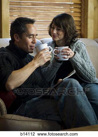 Stock Image - Man and woman on sofa with mugs smiling. Fotosearch