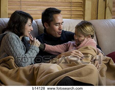 Stock Photo - Man and woman snuggling with young girl on sofa with blanket. Fotosearch
